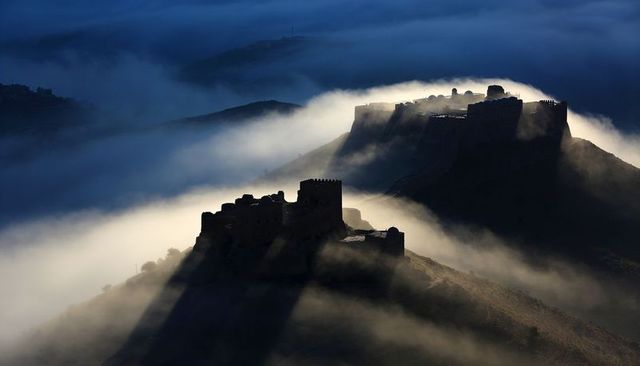 Ruined hilltop forts rising through morning mist, casting long sunlit shadows across valley