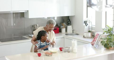 African american grandmother guiding granddaughter mixing batter in bright modern kitchen