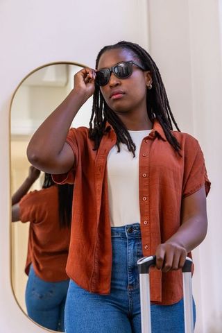 African woman adjusting sunglasses and holding rolling suitcase by mirror, stylish traveler