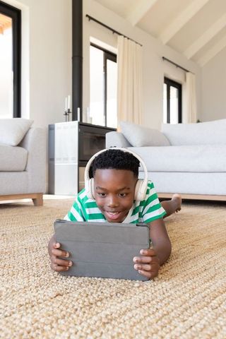 African american boy lying on rug using tablet wearing headphones in sunlit living room