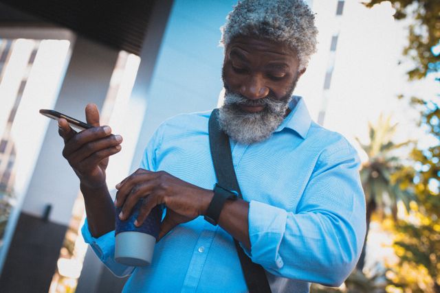 African american senior man standing on street holding coffee using smartphone and smartwatch. digital nomad on the go in the city.