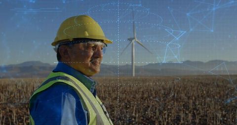 Mature engineer surveying wind turbine at rural windfarm wearing hard hat and hi-vis vest