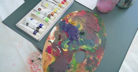 Paint-splattered wooden artist palette with thick impasto colors and tubes on teal table