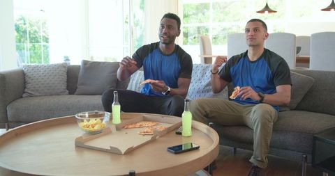 Diverse men watching game eating pizza on couch wearing athletic shirts