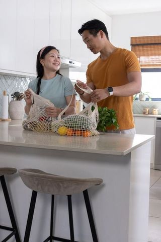Couple unpacking reusable mesh grocery bags with fresh produce on sunlit modern kitchen island