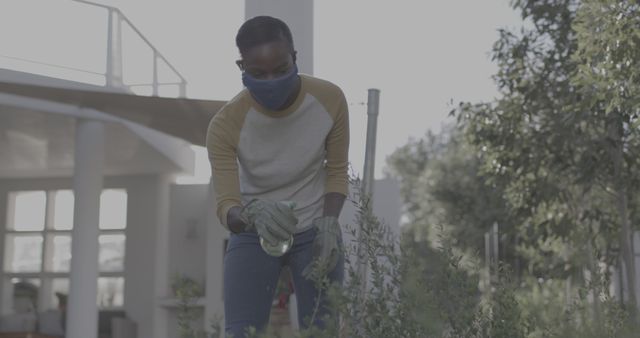 Woman wearing a mask and gloves is spraying plants in her garden. She appears focused on maintaining her garden while practicing safety and hygiene. This image can be used for topics related to home gardening, outdoor activities, safety measures, healthy living, and environmental care.