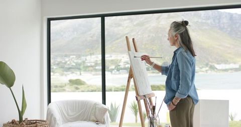 Senior woman painting at easel in sunlit coastal studio with panoramic bay view