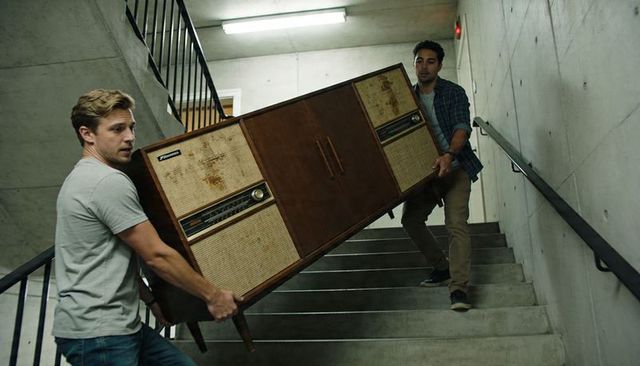 Two men carrying heavy midcentury wooden stereo console down concrete stairwell, teamwork