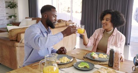 Black couple toasting with orange juice at cozy sunlit breakfast table, avocado toast