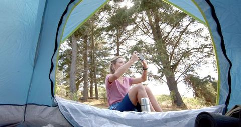 a young woman is sitting inside a tent, capturing moments on her smartphone during a camping trip. she is surrounded by lush green trees, enjoying the tranquil outdoor environment. this scene embodies the connectivity between modern technology and nature adventure. ideal for content related to outdoor activities, camping gear, travel blogs, or promoting adventurous lifestyles.