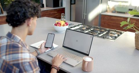 Man working remotely at kitchen island holding smartphone typing on laptop with coffee mug