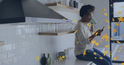 Sipping coffee and texting on smartphone in bright minimalist kitchen with gold stars