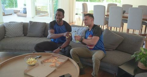Diverse friends clinking bottles on sofa during casual game day with pizza and chips at home