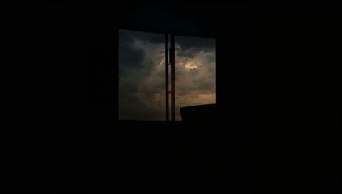 Square window framing stormy dusk sky over silhouetted room with mullion, latch and sofa