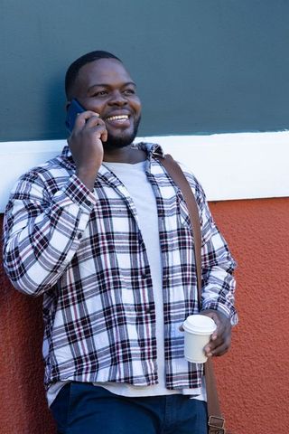 Smiling african american man talking on smartphone holding takeaway coffee leaning on painted wall
