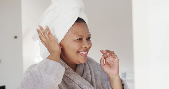 An African woman with a towel wrapped on her head is smiling and touching the towel in a relaxed setting. This is perfect for promoting self-care, skincare products, morning routines, beauty and wellness, or hygiene products. Businesses can use it for social media promotions, website banners, or advertorial content related to health and beauty.