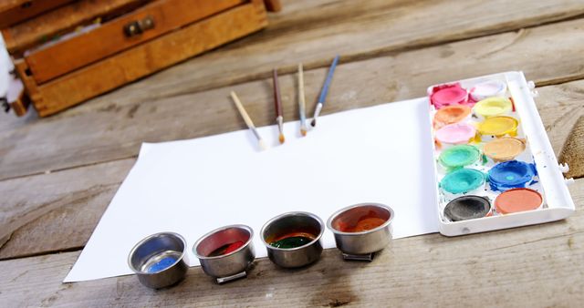 A set of watercolor paints, brushes, and small cups of water are ready for an artist to begin painting, with copy space. Creativity and artistry are suggested by the arrangement of painting supplies on a rustic wooden surface.