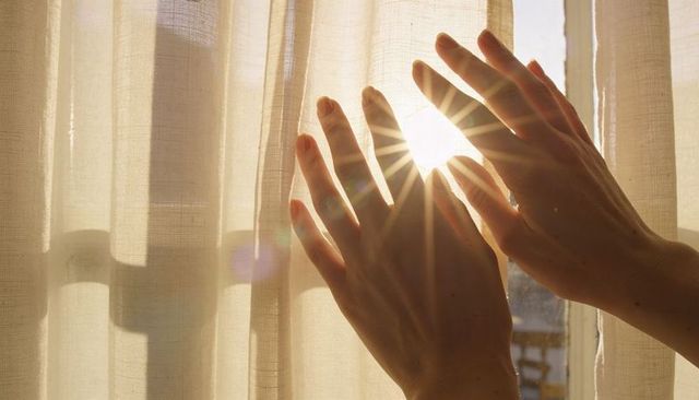 Adult hands parting sheer curtain letting sunlight starburst through window at golden hour
