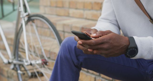 Close-up of person sitting on steps next to bicycle using smartphone. Person wearing a smart watch, suggesting an interest in tech or sports outdoors. Perfect for themes such as modern technology, urban lifestyle, outdoor activities, commuting and healthy living.