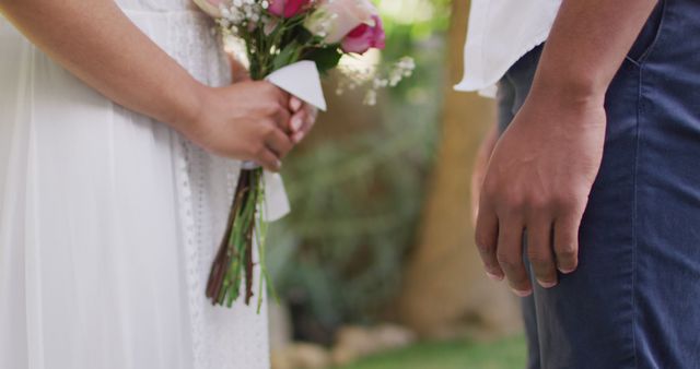 Couple holding hands and bridal bouquet during an outdoor wedding ceremony. Ideal for representing romantic occasions, engagements, or marriage-related content. Suitable for wedding planning websites, romantic greeting cards, and social media posts about weddings.