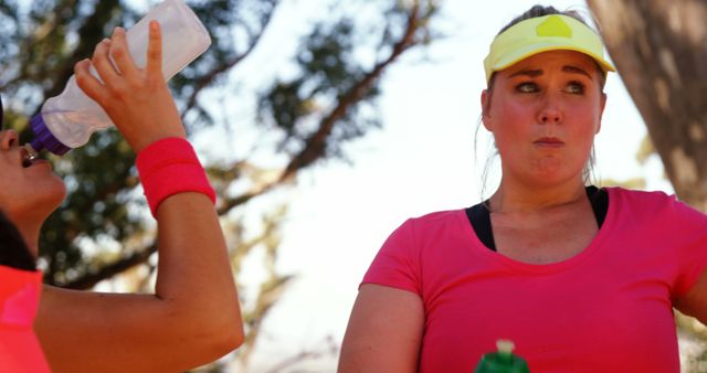 Two young women taking a break to hydrate while engaging in an outdoor workout, wearing bright sportswear and standing near some trees. This visual is useful for fitness and exercise content, promoting the importance of hydration, and showcasing sporty apparel.