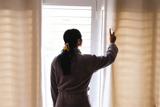 This image depicts a woman in a bathrobe opening curtains in the morning, viewed from the rear. It is ideal for use in lifestyle blogs, articles about morning routines, home living, relaxation, and domestic life. The natural light and casual setting make it suitable for content related to everyday life and home comfort.