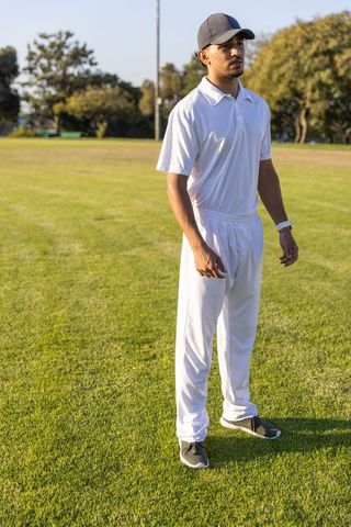 Young indian cricketer standing on sunlit field wearing white kit and dark cap