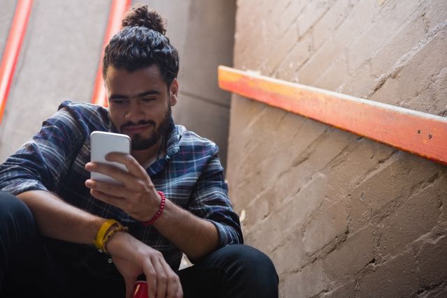 Young man with beard and plaid shirt using mobile phone while sitting on staircase in urban environment. Ideal for themes related to modern lifestyle, technology, communication, and urban living.