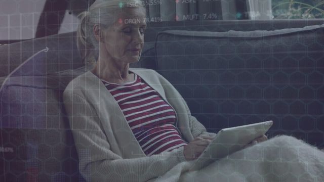 Senior woman comfortably sitting on a couch and using a tablet, with a digital overlay of financial data and charts. Suitable for illustrating fintech concepts, senior citizens using technology, financial analysis while relaxing, or at-home digital interface use.