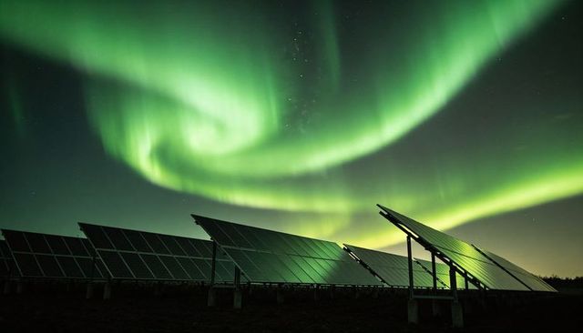 Aurora borealis swirling above solar panels reflecting green glow over night solar farm
