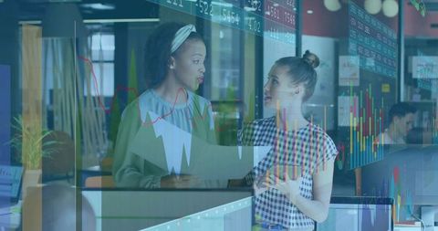 Collaborating women analyzing market charts through glass partition in modern open office