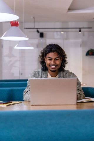 Young indian man working on laptop in modern co-working space with teal booth seating