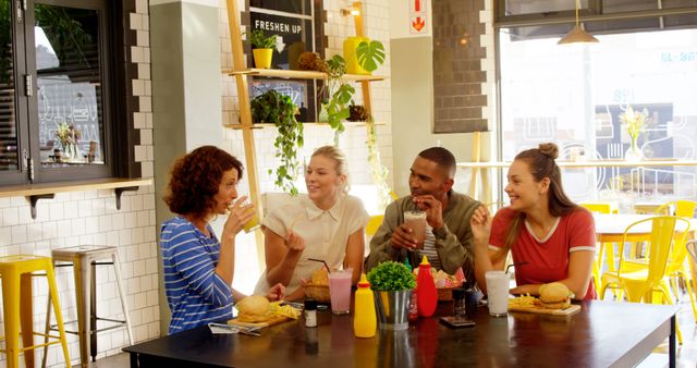 Group of four friends sitting together at a table in a modern, brightly lit cafe. They are smiling and chatting while enjoying their drinks and food. This image is ideal for promoting social gatherings, cafes, restaurant advertisements, and the importance of friendship and togetherness.