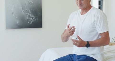 Senior man applying moisturizer to hands while sitting on bed in minimalist bedroom