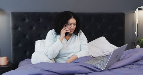 Woman Working from Bed in Pajamas, Talking on Phone and Checking Laptop for Remote Work