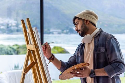 Indian artist painting at easel in sunlit waterfront studio wearing beanie and headphones