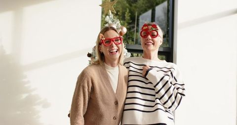 Two women laughing in festive novelty glasses by christmas tree in bright sunlit room