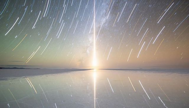 Vertical light beam reflecting on glossy salt flat with milky way star trails, long exposure