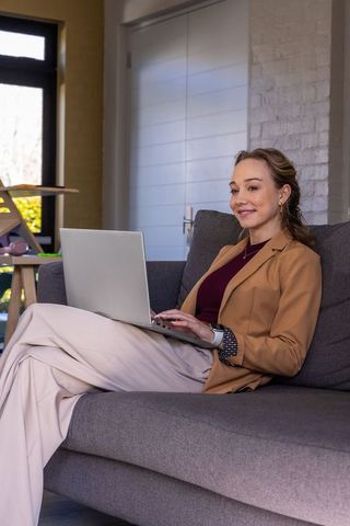 Smiling professional woman working remotely on laptop at home on gray sofa using smartwatch