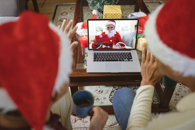 Diverse couple wearing Santa hats connecting with a man dressed as Santa Claus through a laptop video call. Celebrating Christmas virtually with festive decorations, gifts, and a warm holiday atmosphere. Ideal for themes of online communication, holiday celebrations, and connecting across distances during festive seasons.