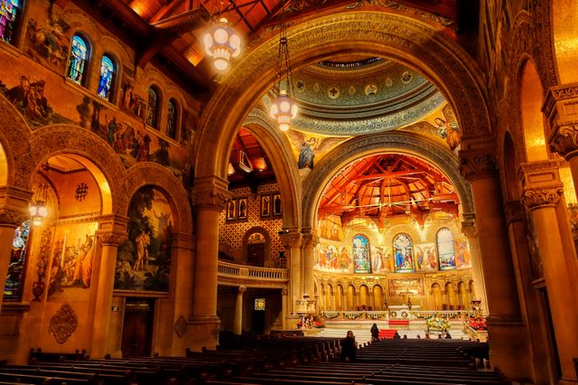 Photo captures breathtaking interior of an ornate Anglican cathedral. Rich decorative elements include stained glass windows, arched ceilings, and detailed frescoes. Scene suggests reverence and tranquility, with an empty pew area overlooking the illuminated altar. Perfect for use in articles about religious architecture, historic structures, and interior design inspirations.