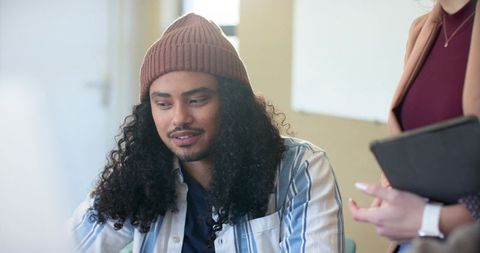 Curly-haired man wearing beanie and colleague reviewing tablet during collaborative office meeting