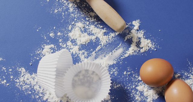 Flour and eggs arranged with a rolling pin and cupcake liners on a blue surface. Ideal for use in cooking blogs, recipe websites, culinary magazines, or menu designs. Conveys a sense of baking preparation and creativity in the kitchen.
