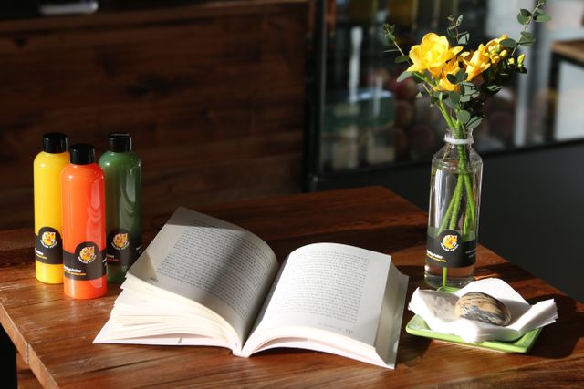 Open book alongside vibrant colored bottles and a vase with yellow flowers on wooden table, suggesting a relaxed and studious ambiance. Ideal for promoting reading campaigns, cafe advertisements, lifestyle blogs, and educational content.