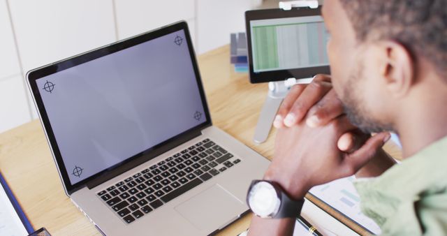 African American man sitting at desk in office environment working on laptop with hands clasped, displaying focus and productivity. Another screen visible in background indicating multitasking. Ideal for use in content related to business, technology, remote work, and professional environments.