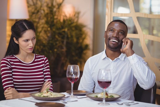 Man talking on mobile phone while woman looks bored in restaurant. Ideal for illustrating relationship issues, communication problems, and the impact of technology on social interactions. Useful for articles, blogs, and advertisements related to dating, relationships, and modern communication challenges.