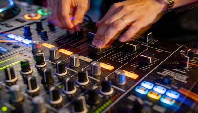 Dj mixing hands adjusting faders and knobs on backlit mixer with neon led meters