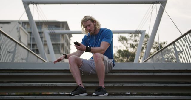 Caucasian man with earphones using smartphone sitting on stairs in city. Sports, fitness, healthy living, communication and outdoor activities, unaltered.