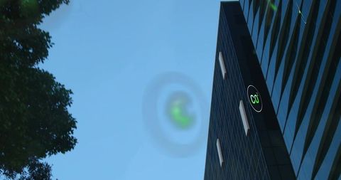 Low-angle glass skyscraper soaring beside tree, green C0 logo glowing under blue-sky halo