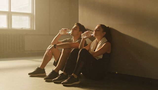 Sunlit fitness couple hydrating and resting after intense training by studio wall, towel